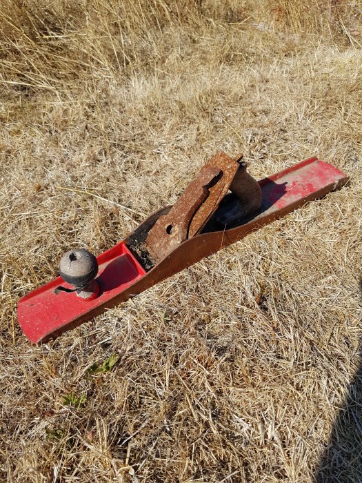 Gammalt hyvelverktyg i rost och rött liggande på torrt gräs.