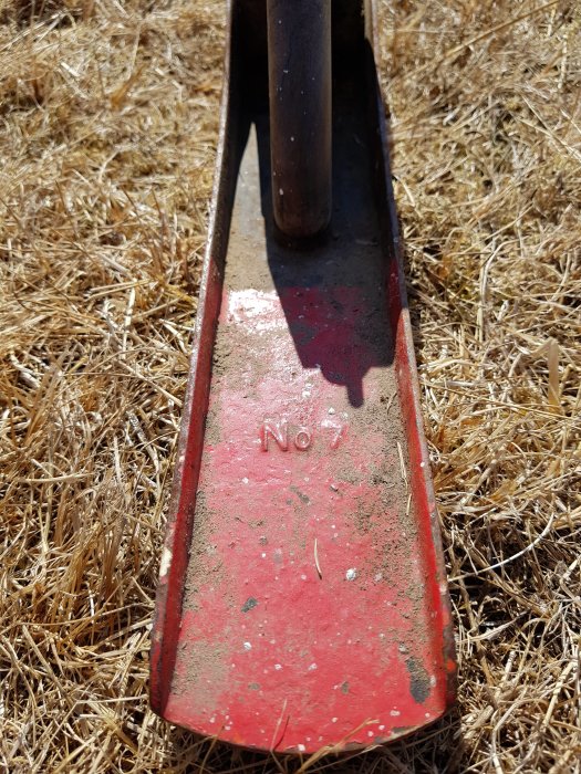 Sliten röd spade med texten 'No 7' på bladet, mot en bakgrund av torrt gräs.