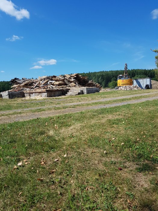 En hög med rivningsvirke och en grävmaskin bredvid containrar på en byggarbetsplats.