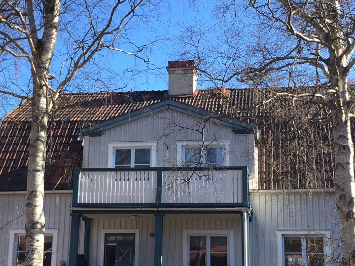 Hus med slitet tak före renovering, omgivet av träd och blå himmel i bakgrunden.