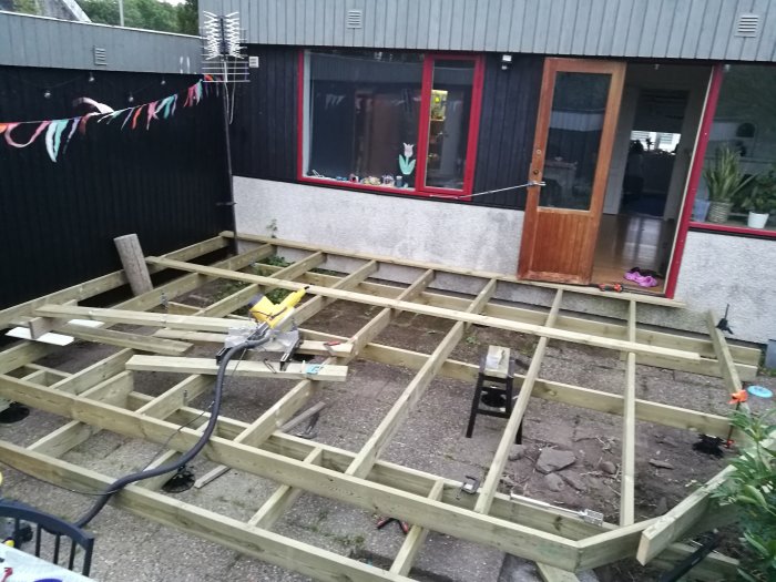 Pågående konstruktion av träregelverk för en altan utanför ett hus med röda och svarta detaljer i skymningen.