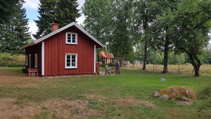 Röd stuga med vita knutar och svensk flagga i en lantlig miljö med kanin framför.