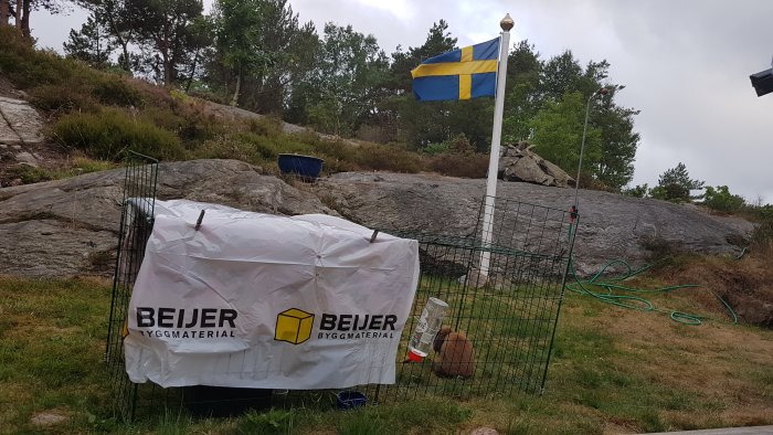 Vädurskanin i bur utomhus bredvid svensk flagga och kompostgaller med Beijer-logotyp.