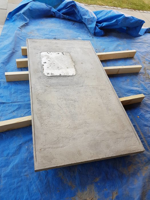 Nyutgjuten betongskiva som inte är helt plan, vilandes på träreglar på en blå presenning.