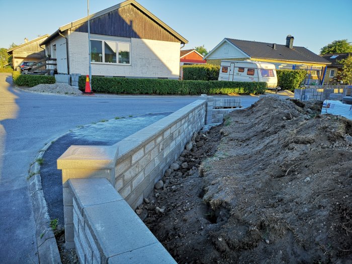 Delvis byggd mur och jordmassor vid ett hus, material och utgrävningsarbete syns.