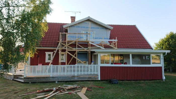 Rött hus med tillfälliga byggnadsställningar och skuggande björk i soligt väder.