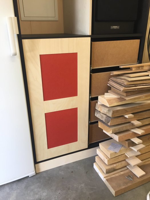 En del av ett högtalarskåp med röda paneler bredvid en hög med träplankor i en verkstad.