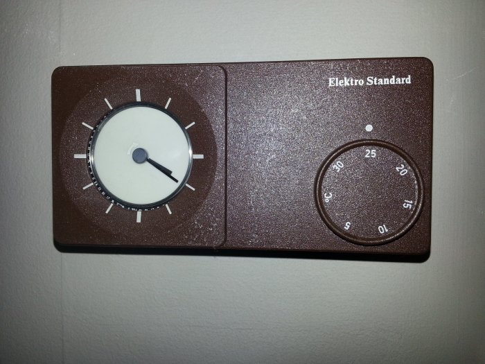 Gammal brun termostat med vridknappar på vit vägg, märkt "Elektro Standard".
