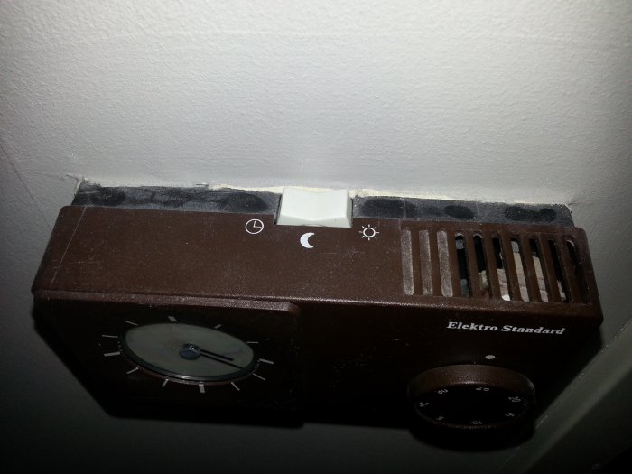 Gammal brun termostat på vägg med vred och reglage, märkt "Elektro Standard".