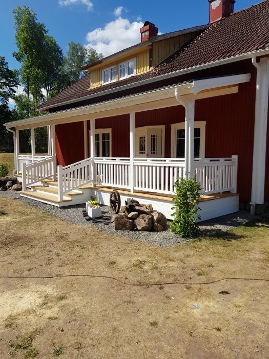 Renoverat rött hus med vit veranda och gula detaljer under soligt väder.