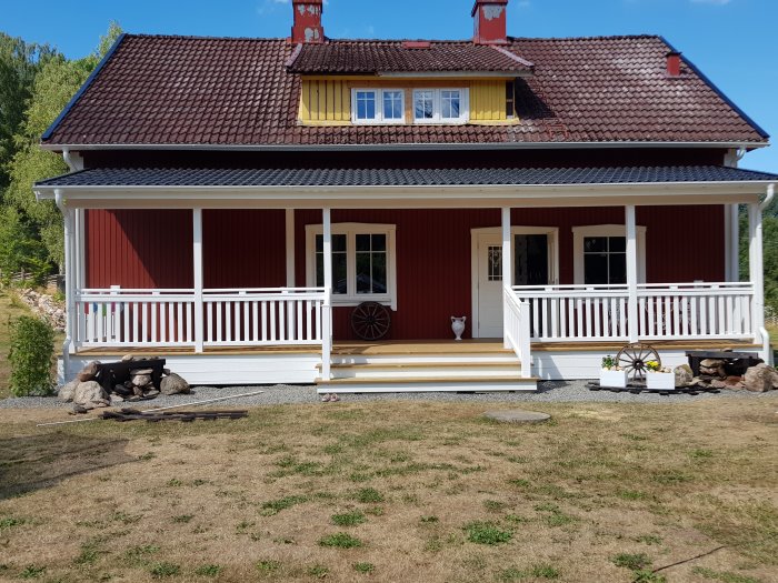 Renoverat rött hus med vit veranda och takstolar, nästan färdigt byggprojekt i sommarlandskap.