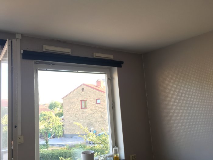 Åpen vindu i rommet med murhus og grønt landskap utenfor, ventilasjonsgitter over vinduet og enkel innredning rundt.
