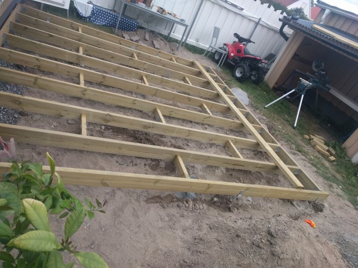 Nygjuten plintgrund med regelverk av trä för ett däck, bredvid en röd trädgårdstraktor och såg på en grusgång.