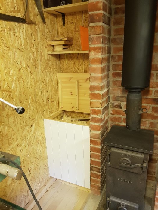 En snickarbod med ett nysnickrat grunt skåp med skjutluckor bredvid en kamin och en vedlår under hyllor.