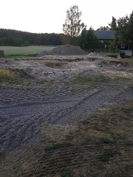 Tom byggplats med grushögar och spår av borttagen grund och murstock, omgiven av grönska.