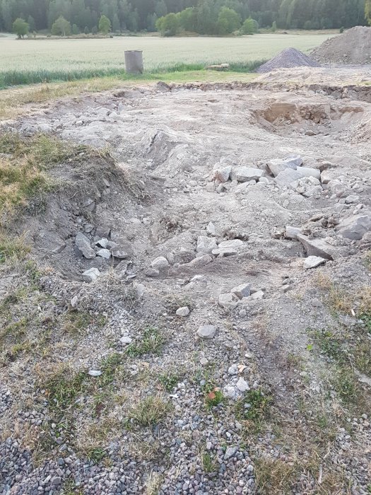 En raserad grund med spridda stenar och grus på en byggarbetsplats, med åkrar i bakgrunden.