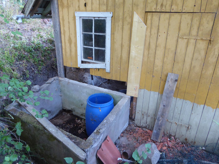 Hörn av ett gulmålat hus med fönster ovanför en förberedd gjutform i betong, samt armering och stödmaterial.