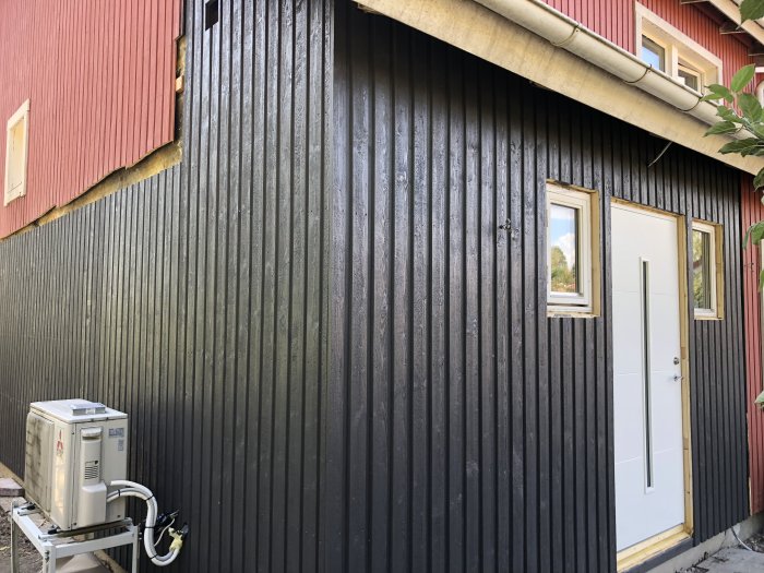 Ny målad fasad och entrédörr i vitt på hus med röd träpanel, nära golfbana på varm dag.