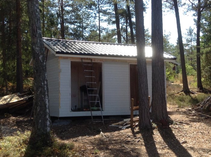 Nybyggd stuga med vit panel nästan klar, omgiven av skog och byggmaterial på marken.