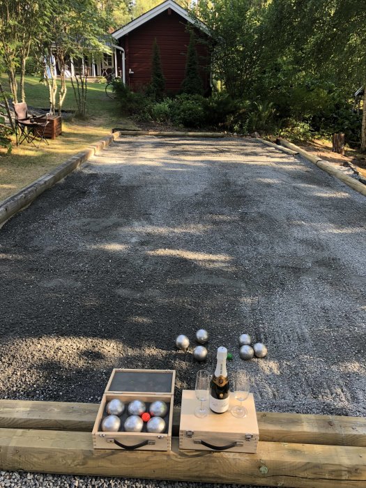 Boule-bana med grus, bouleklot och champagne vid en trädgårdsfest, röd stuga i bakgrunden.