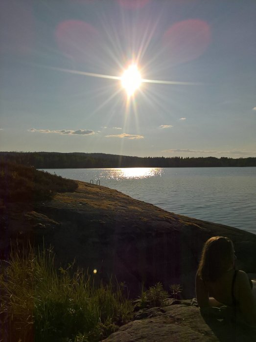 Kvällssol över en sjö, sett från en klippa med person i förgrunden och badstege i bakgrunden.