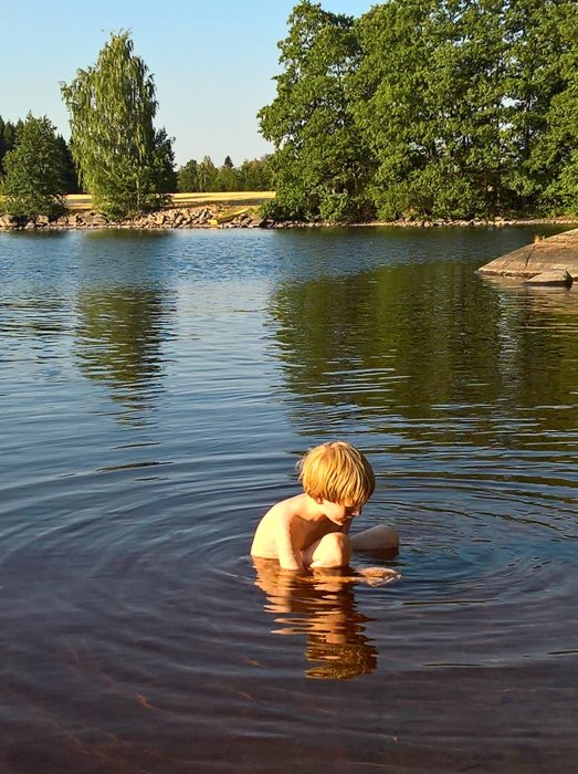 Barn badar i sjö med gröna träd och stenstrand i bakgrunden.