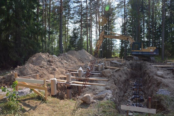 Byggarbetsplats med nyligen gjutna betongplintar och återfylld jord, grävmaskin och skog i bakgrunden.