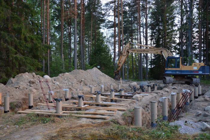 Byggplats med nygjutna betongplintar och en grävmaskin i bakgrunden omgiven av skog.