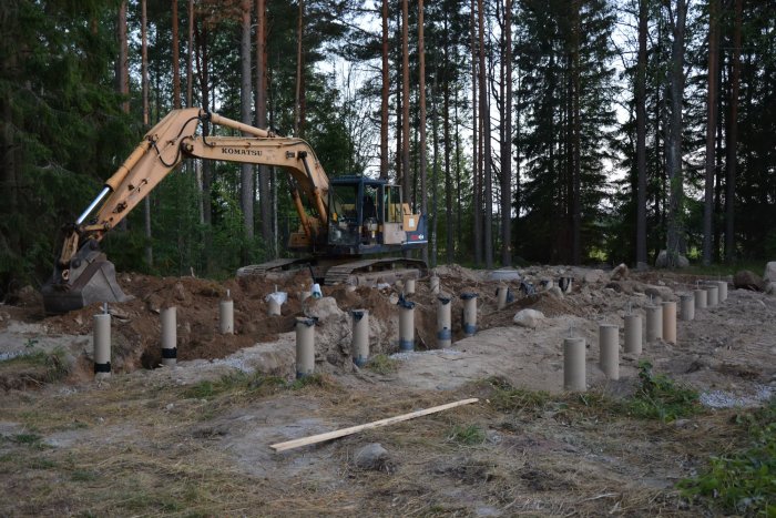 Grävmaskin vid byggarbetsplats med gjutna betongplintar i skogsmiljö och uppgrävd mark.