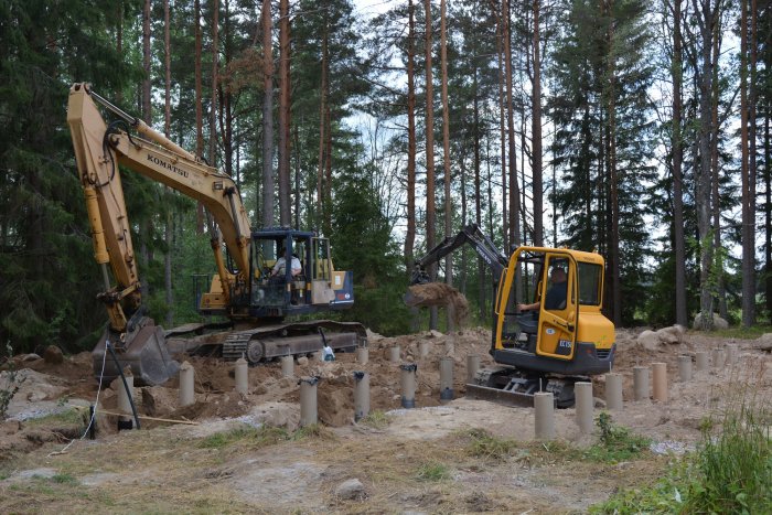 Byggarbetsplats med nyligen gjutna betongplintar och grävmaskiner som arbetar med återfyllning.