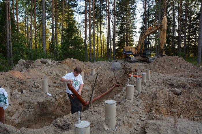 Byggprojekt där betongplintar och avloppsrör installeras manuellt med grävmaskin i bakgrunden.