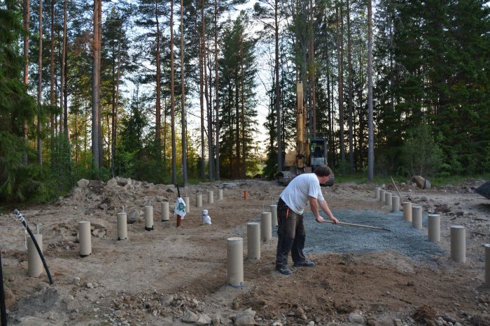 Grundläggningsarbete med person som jämnar ut grus vid plintar i skogsmiljö, byggarbetsplats förberedd för fuktskydd.