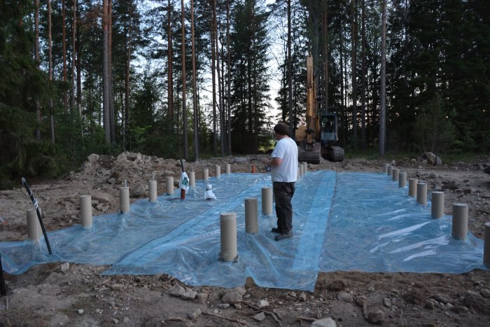 Underlagsarbete för husgrund med plastfolie och betongplintar i skogsmiljö, en person och en grävmaskin syns.