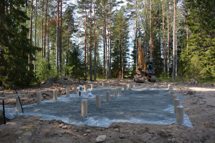 Underlagsarbete med husgrunden: plastduk, grus och betongplintar i skogsmiljö redo för limträbalkar.