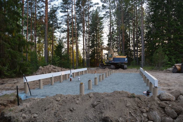 Nygrundlagd byggplats med plintar och limträbalkar i skogsmiljö, grusunderlag, och en grävmaskin i bakgrunden.