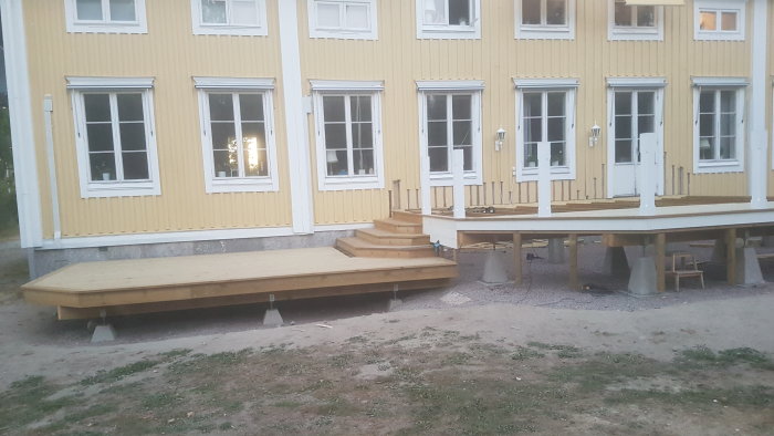 Översiktsbild av ett gult hus med vita fönster och en nybyggd trätrappa och terrass i förgrunden.