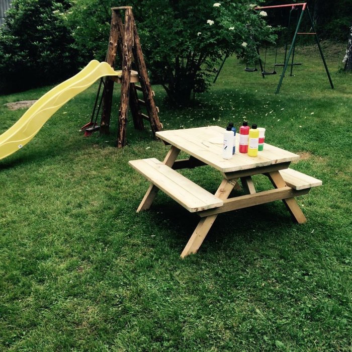 Hemmagjord picknickbänk av trallvirke i trädgården med färgfläckar och en rutschkana i bakgrunden.