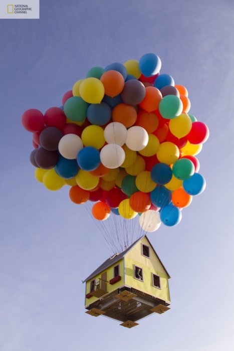 Hus som svävar i luften hållet upp av ett stort knippe färgglada ballonger mot en klarblå himmel.