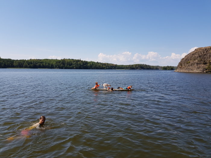 Personer badar och solar på en gammal vindsurfbräda i en sjö, omgiven av natur och klippor.