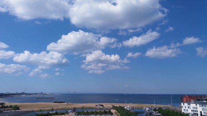 Vy över kustlinje med moln, Öresundsbron i bakgrunden, båtar och byggnader.