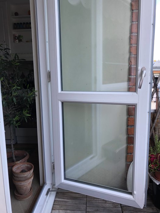 Vit balkongdörr med lås, öppen inåt, med utsikt över en balkong med växter och krukor.