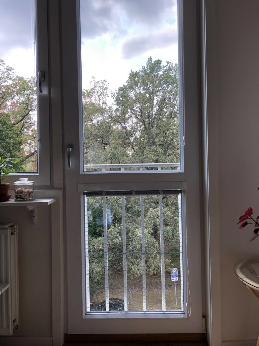 Vy genom en öppen balkongdörr med lås och säkerhetsgaller som vetter mot träd och himmel.