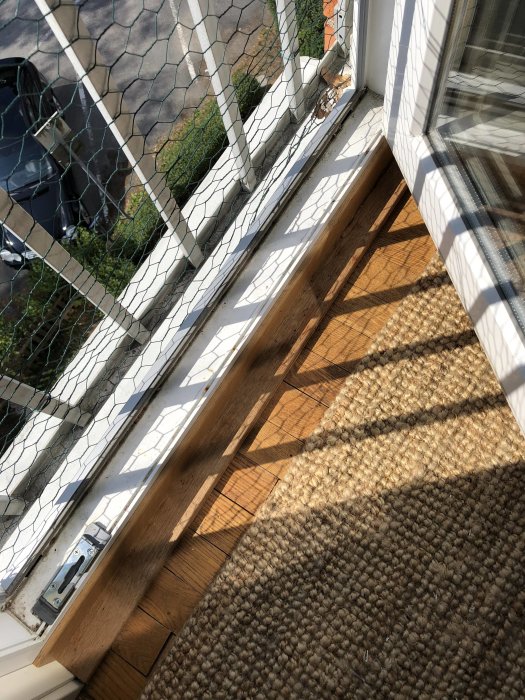 Bild av en öppen balkongdörrs undersida med lås, sett inifrån, med solljus som skapar skuggor på trägolvet.