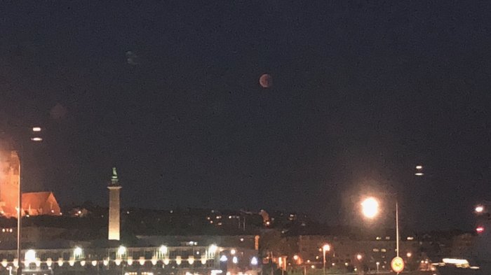 Kvällsutsikt över stadssilhuett med upplysta byggnader och en rödaktig måne på himlen.