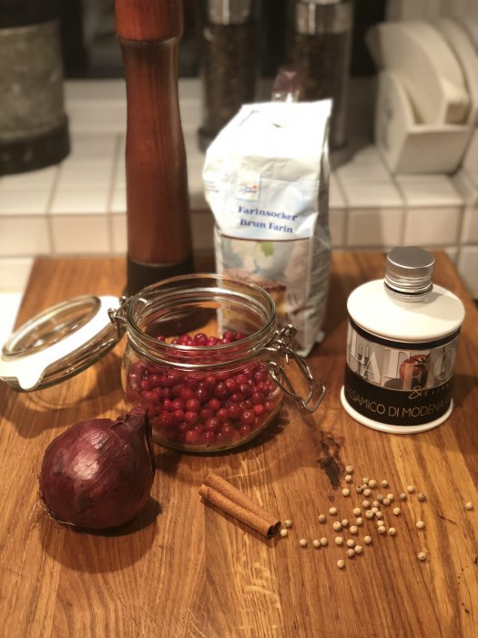 Ingredienser för lingon- och rödlökschutney: lingon i glasburk, brun farinsocker, stor rödlök, kanelstång och vitpepparkorn på träyta.