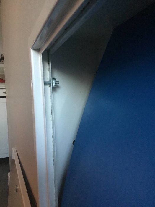 Detaljbild av en vit luckram fäst med maskinskruv i ett skåp med synligt mellanrum mot en blå vägg.