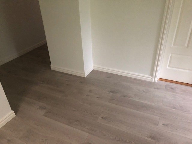 Renoverat rum med nytt laminatgolv, vita väggar, foder och lister vid dörröppning.