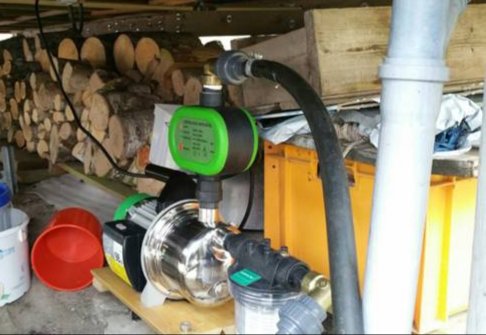 Vattenpump installerad under träbänk med sugslang, kopplingar och tryckvakt, i ett utrymme med vedstaplar.