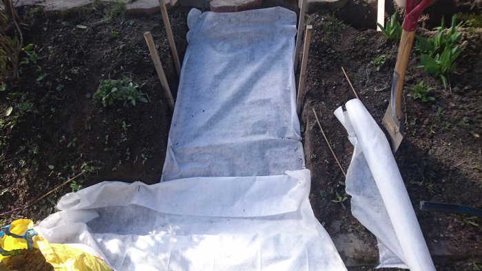 Trädgårdsprojekt med markduk utlagd mellan träpinnar och spade i förgrunden.