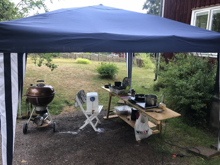 Improviserat utekök under blått tält med grill och kokkärl i en trädgård.